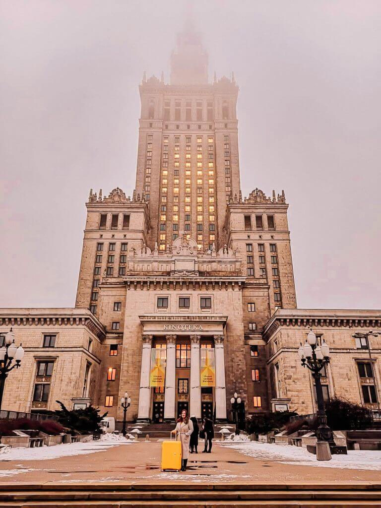 Woman in a beige winter coat holding a yellow suitcase standing in from of The Palace of Science and Nature in Warsaw Poland