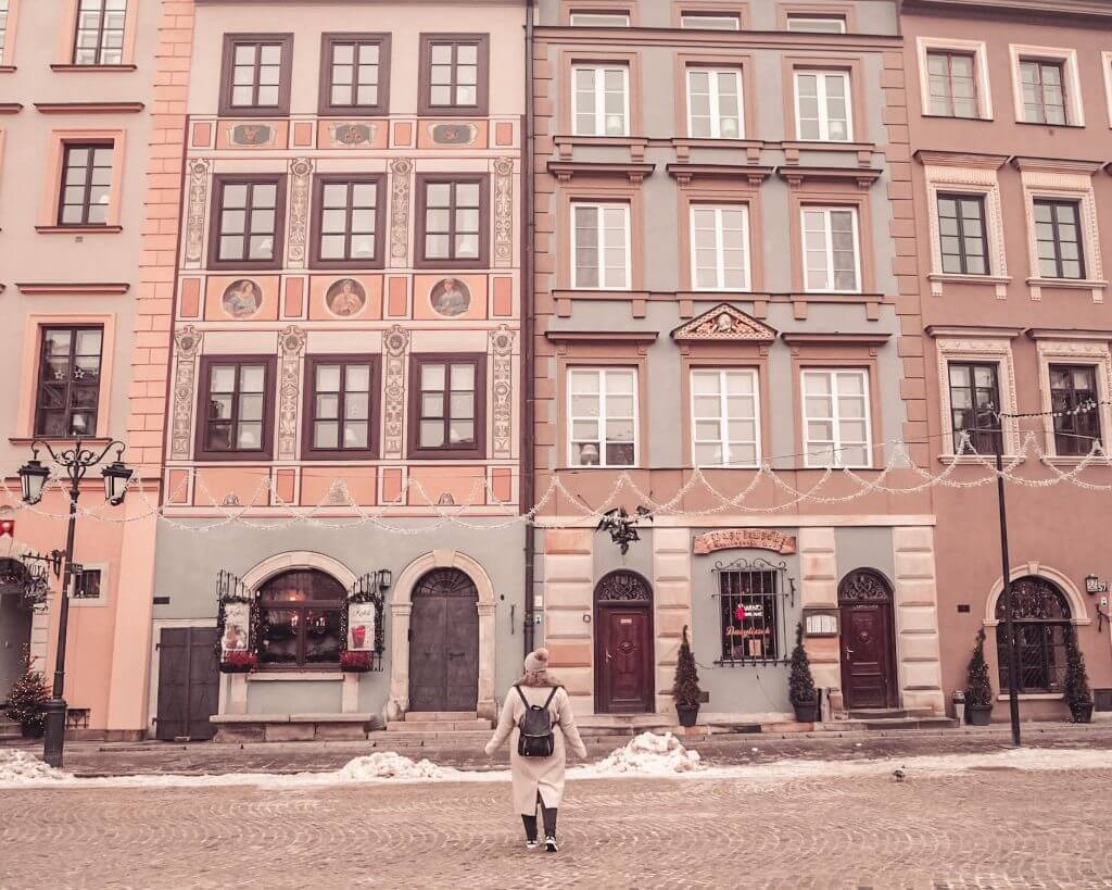 Woman in a beige coat and hat standing in front of the pastel buildings of Warsaw old town square