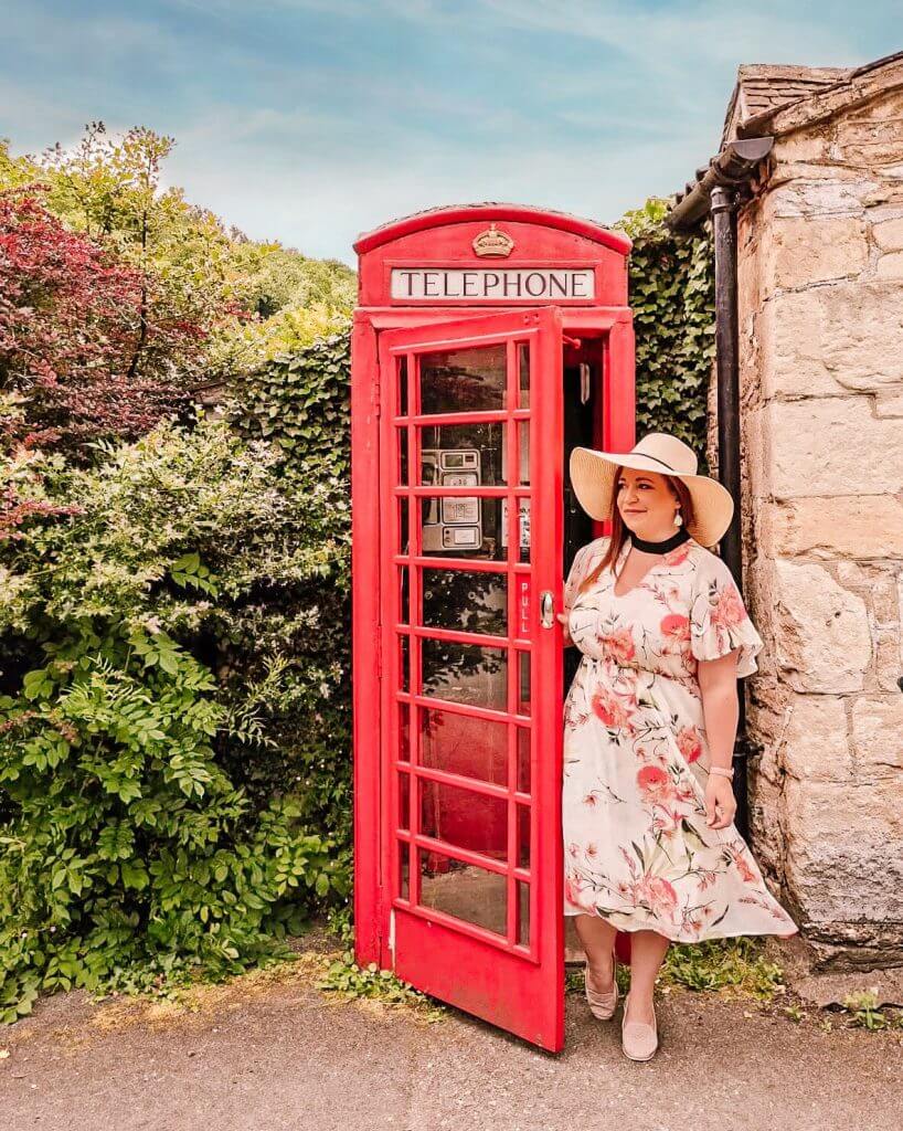 Woman in a floral dress opening the door of a red telephone box in Castle Combe one of the Cotswolds prettiest villages