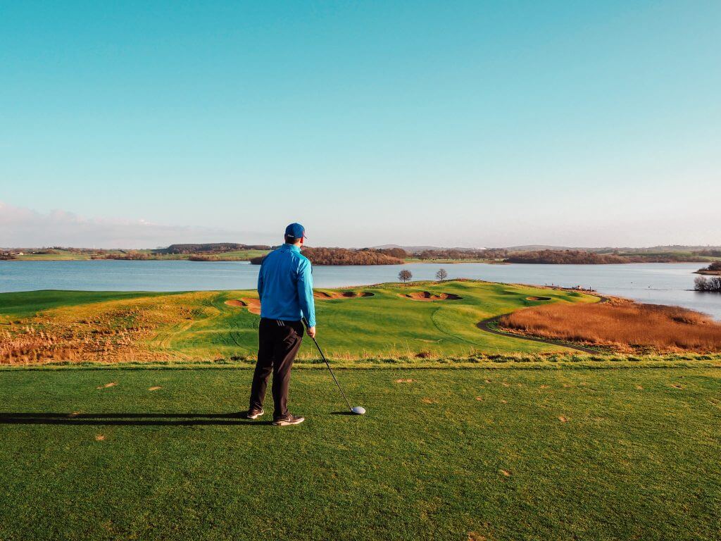 Male golfer in a blue top and black pants holding a golf club and admiring the views from hole 7 at Lough Erne resort.