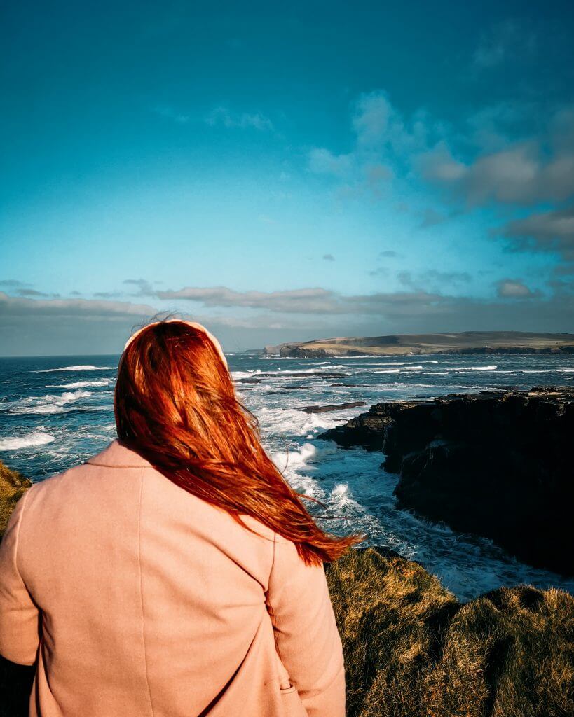 Irish woman with red hair and wearing a pink coat looking out over the Irish coastline.