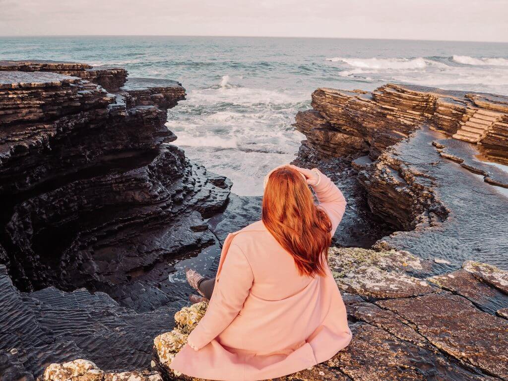Irish woman with red hair wearing a pink coat and looking out to the Atlantic Ocean from a cliff in Ireland.