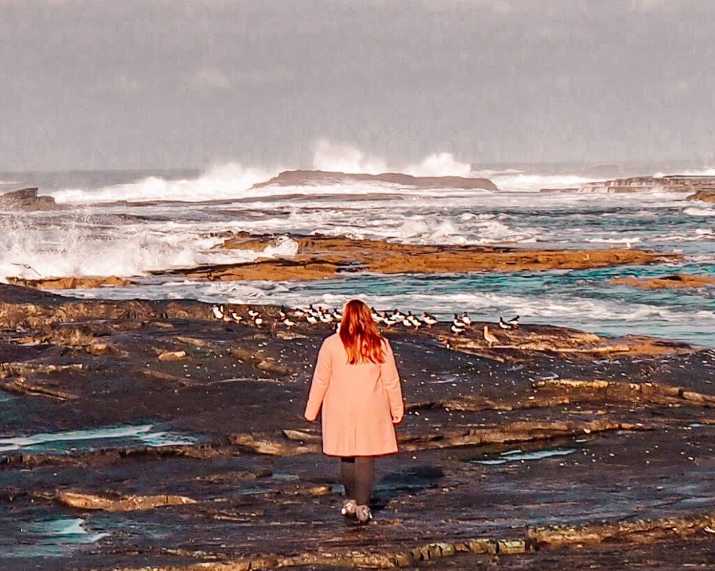Irish woman with red hair wearing a pink coat and walking along a stony beach in Kilkee County Clare Ireland.
