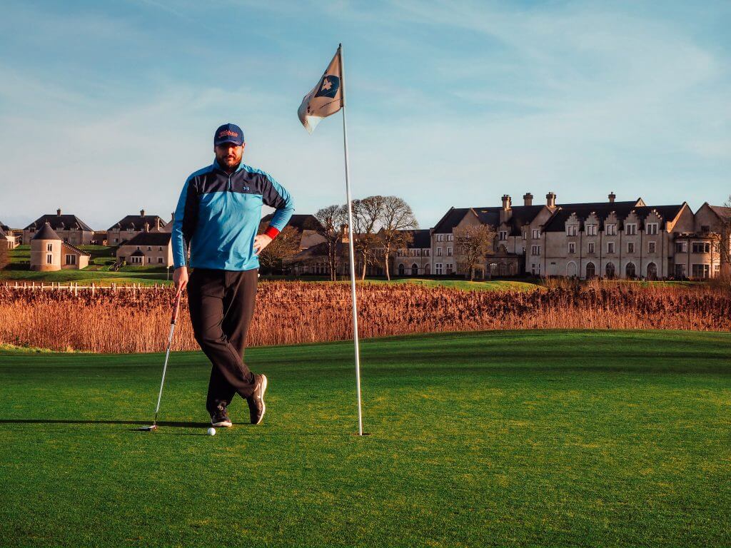 Man in blue top standing beside a golf flag after putting his golf ball