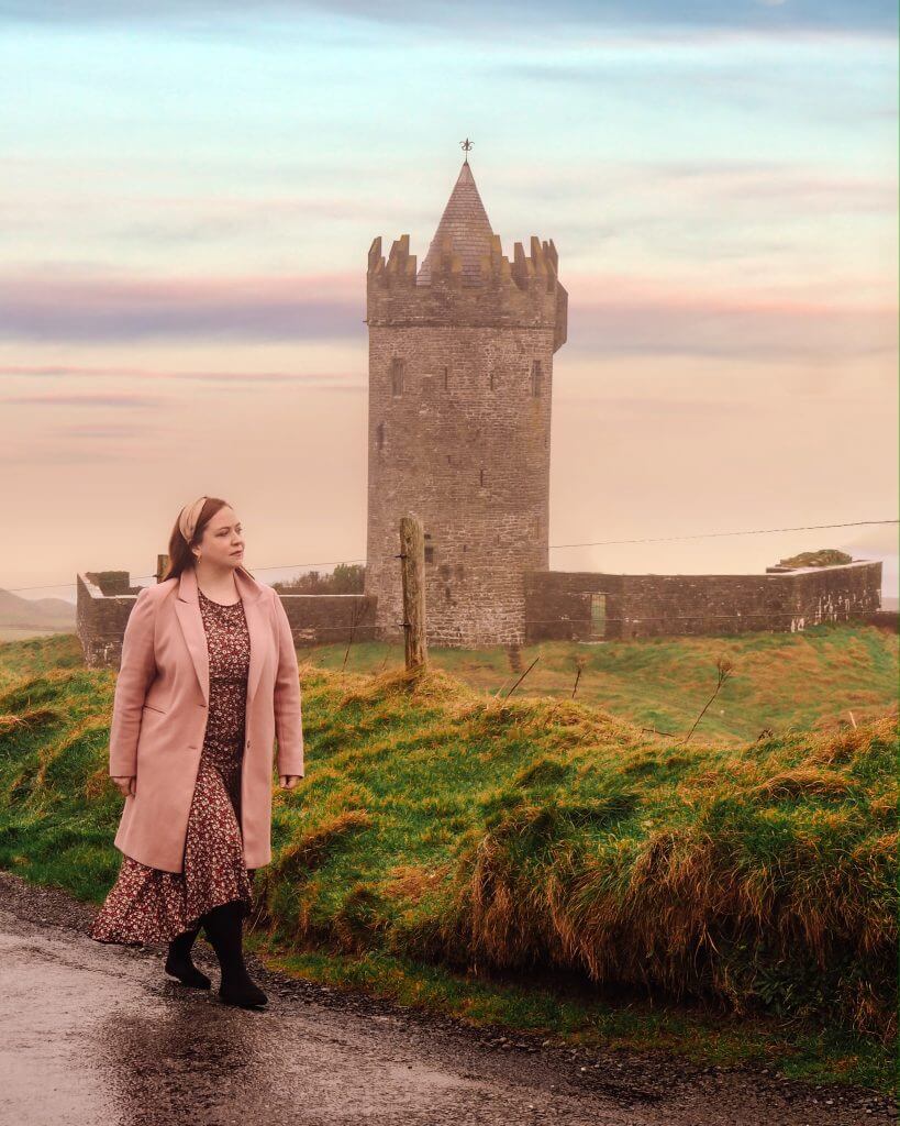 Woman wearing a floral dress and a pink coat walking past a beautiful old tower house in County Clare in Ireland.