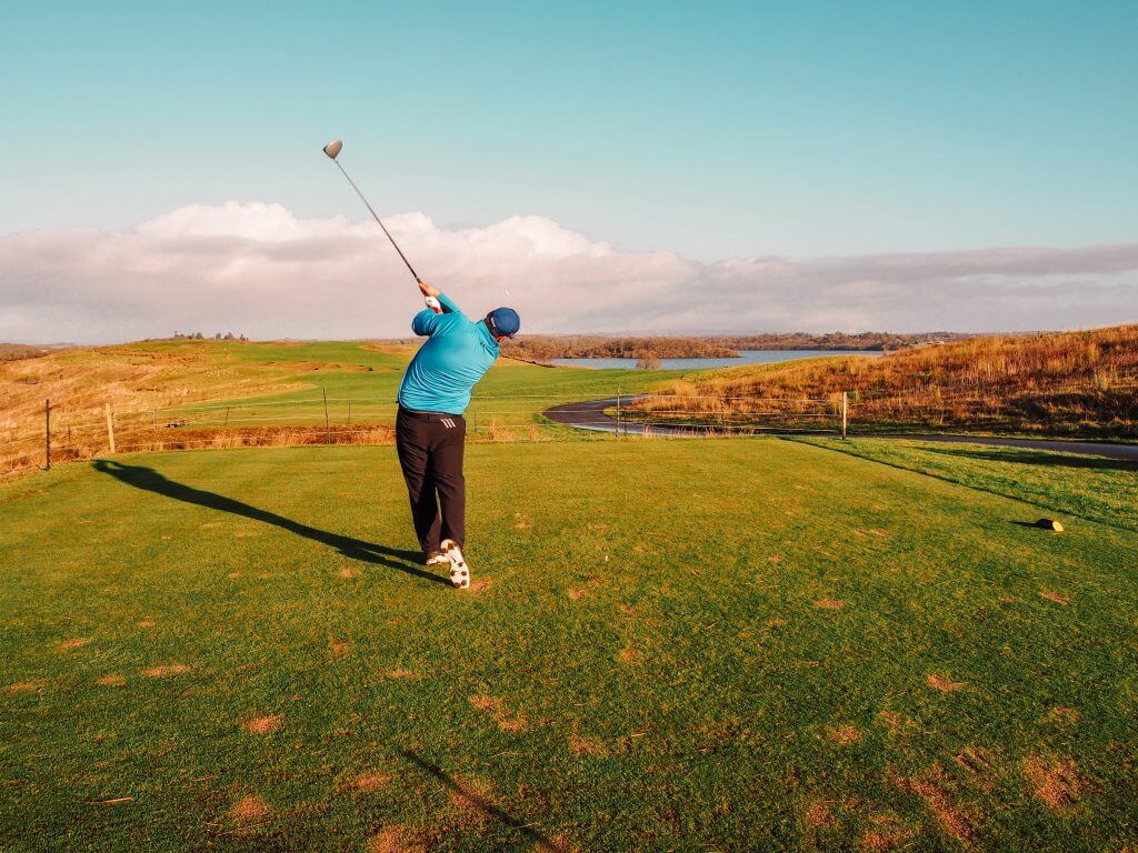 Male golfer in blue top and black trousers swinging a golf iron.