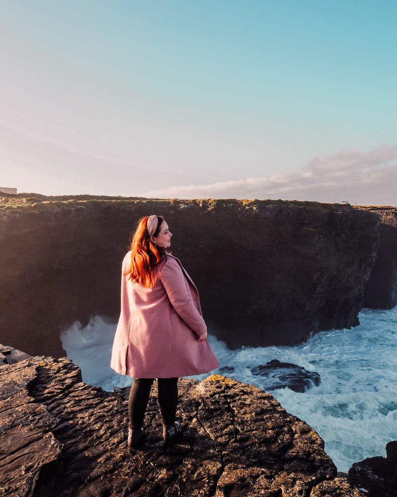 Woman in a pink coat standing at the edge of Kilkee cliffs in County Clare Ireland overlooking the crashing waves of the Atlantic Ocean