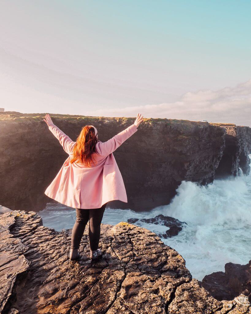 Irish Woman with red hair wearing a pink coat has her hands up in the air as she stands on a clif edge as the waves of the Atlantic Ocean crash below her.