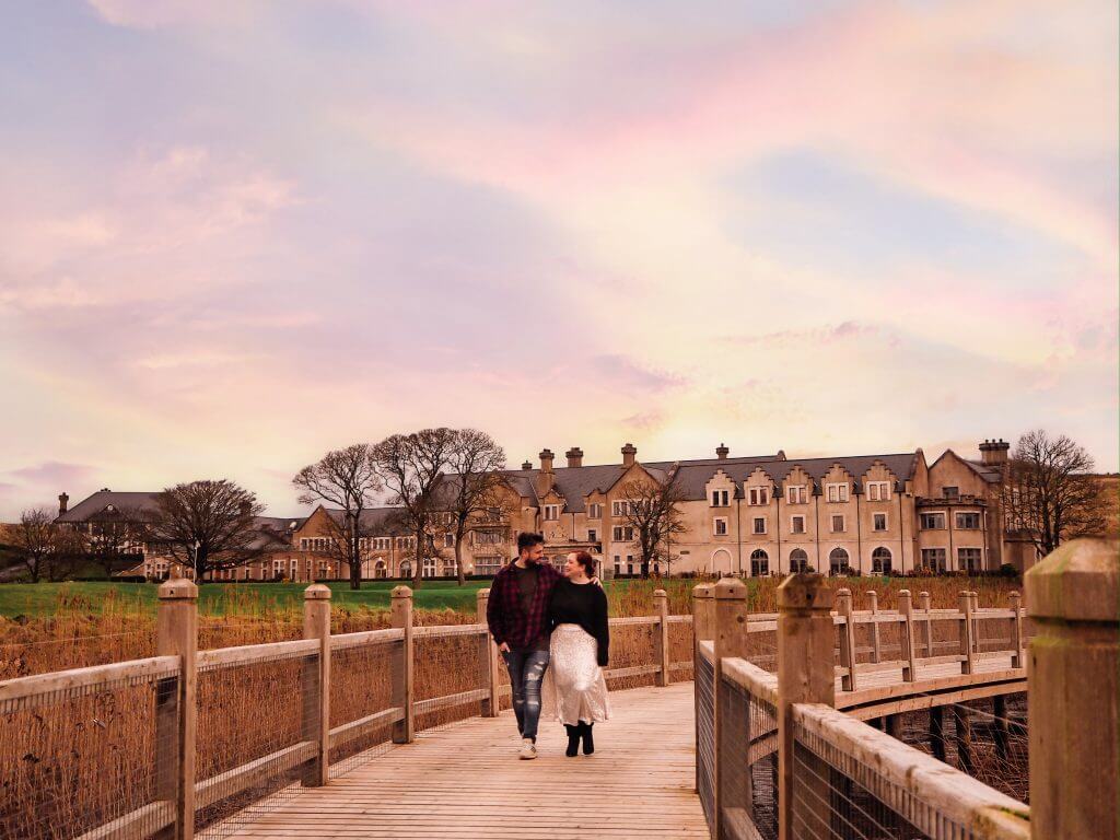 Couple walking hand in hand over a wooden bridge at Lough Erne resort one of Ireland's most romantic hotels under a beautiful sunset.