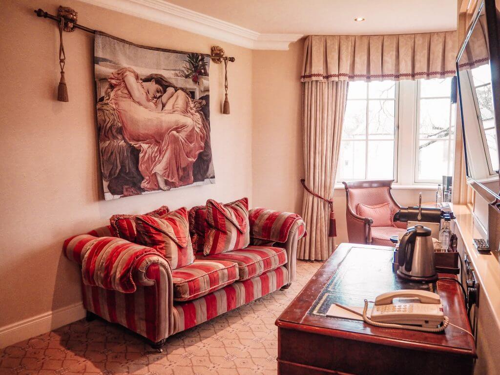 The seating area of the Dovecote suite in Lough Erne resort with a plush velvet red and beige couch and a tapestry of a woman in a pink dress on the wall.