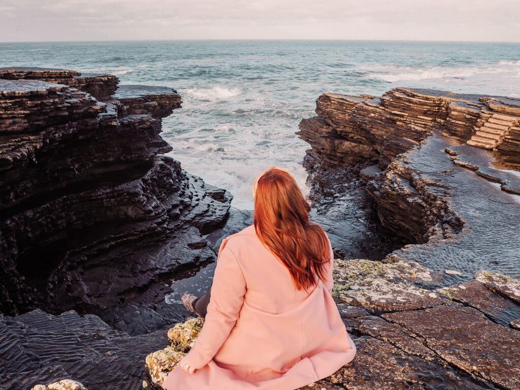 Irish woman with red hair and wearing a pink coat sitting at the cliffs edge at Kilkee Cliffs In County Clare Ireland