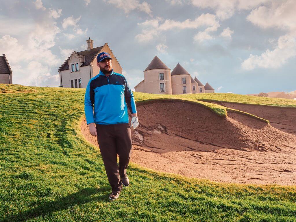 Male golfer in a blue top and black trousers standing in front of a golf bunker at Lough Erne Golf Resort.