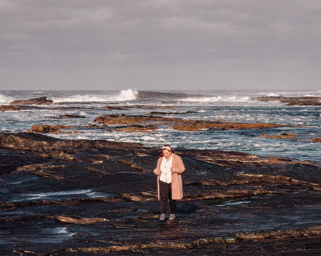 Woman wearing a pink coat and Aran sweater walking along the rocky coastline of Kilkee County Clare in Ireland.