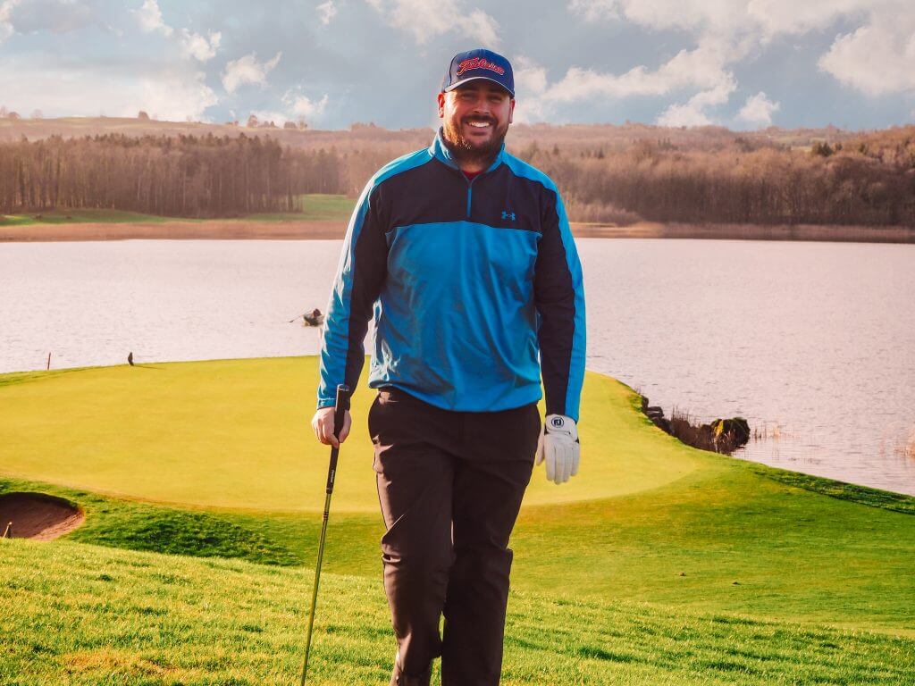 Male golfer in blue top and black trousers smiling and holding a golf club.