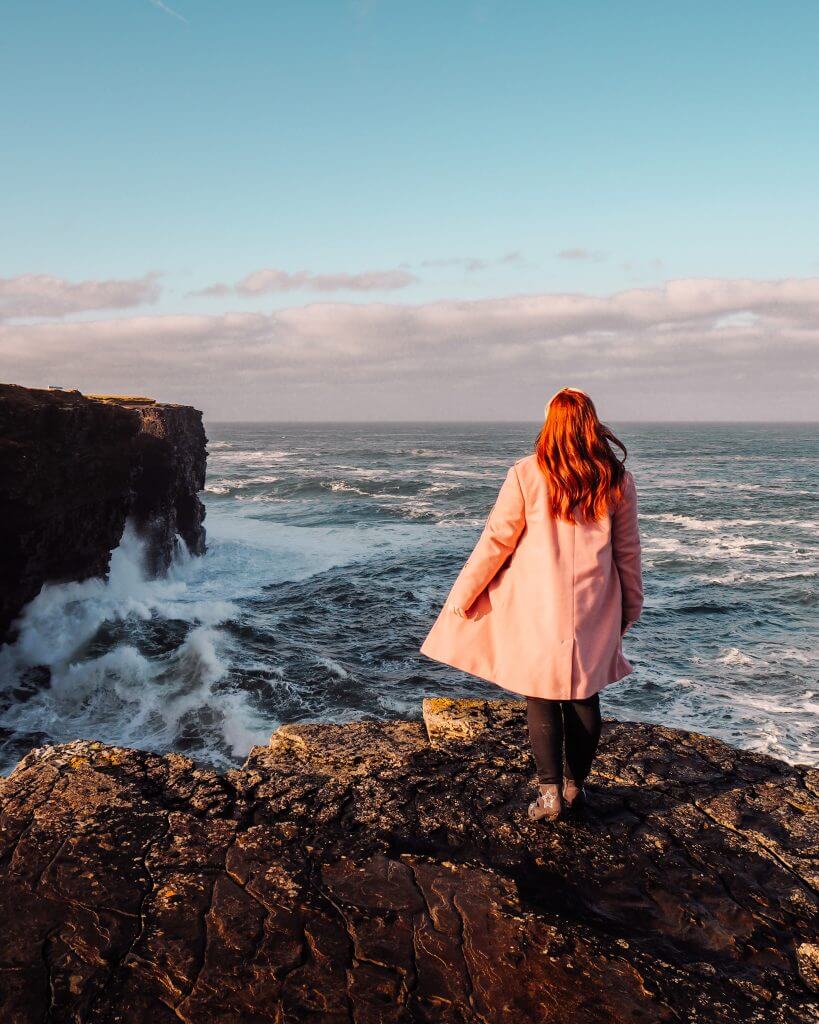 Irish woman with red hair and wearing a pink coat looking out at the wild Atlantic ocean in Ireland.