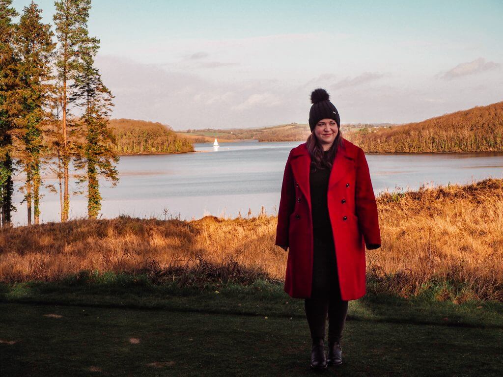 Woman in a red coat overlooking Lough Erne in Northern Ireland as a sailboat sails by.