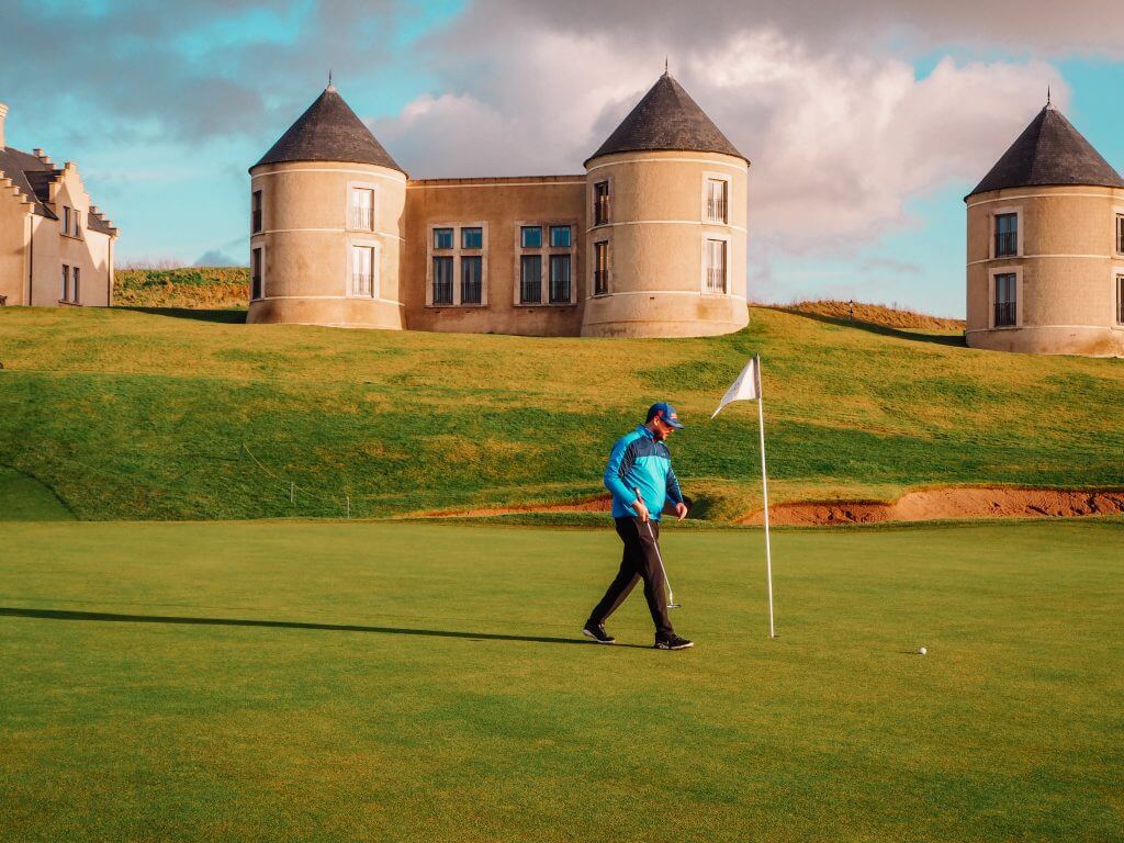 Male golfer in blue top and black trousers walking towards a golf flag.