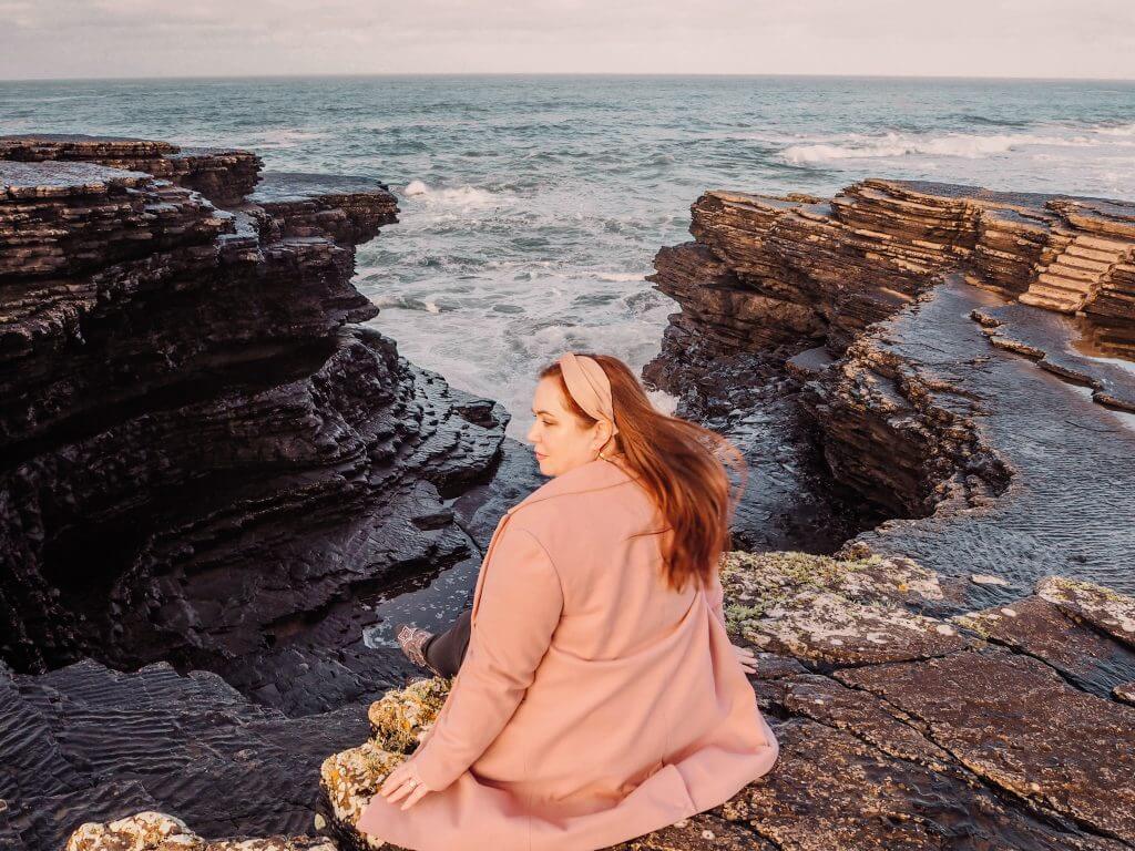 Irish woman with red hair wearing a pink coat and pink hairband sitting on Kilkee Cliff edge overlooking the Atlantic ocean as the waves crash in.