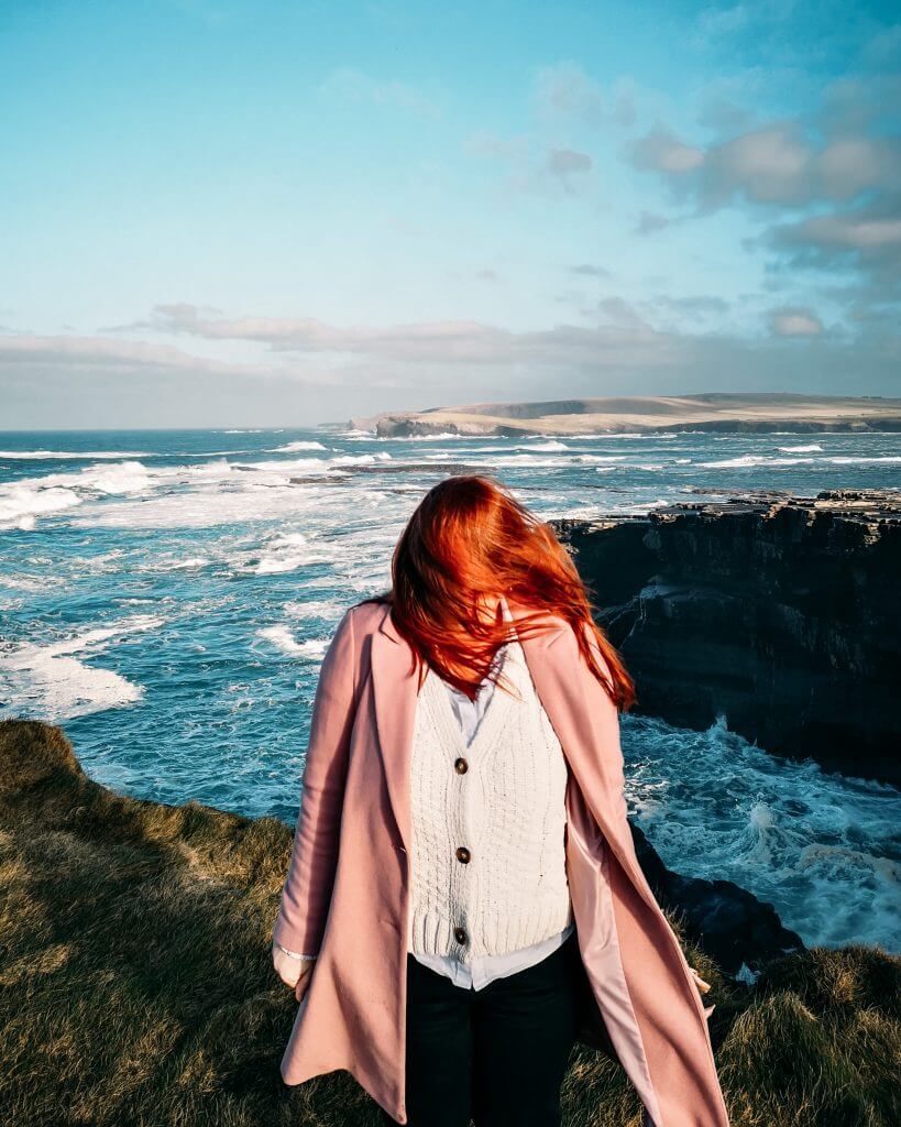 Woman wearing a pink coat and an Aran sweater standing on a cliff edge with the wind blowing her red hair in her face.