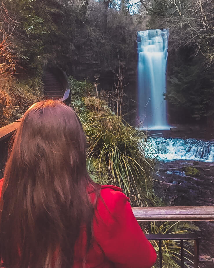 Woman in red coat standing in front of Glencar waterfall in Leitrim Ireland.