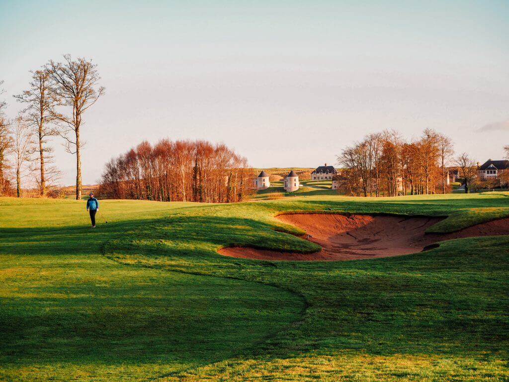 Male golfer in a blue top walking with a golf club near a sand bunker on a golf course in Ireland.