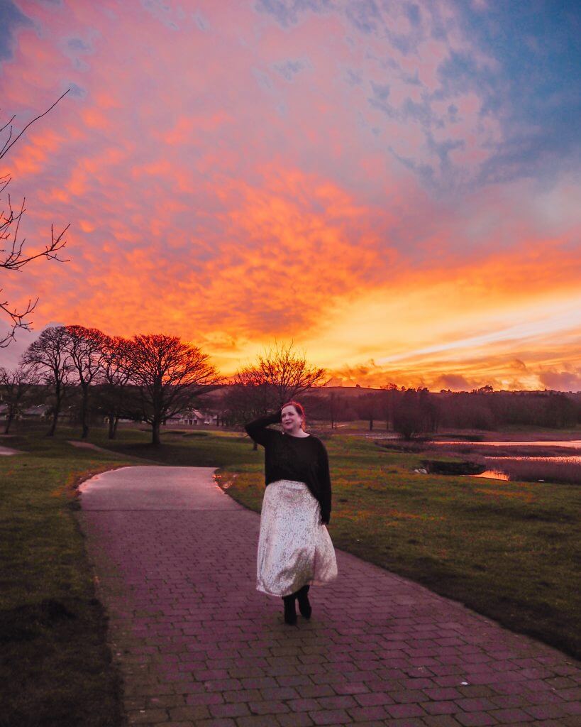 Woman with red hair and wearing a silver sequin skirt walking along the lakeshore of one of Ireland's most luxurious and romantic hotels at sunset.