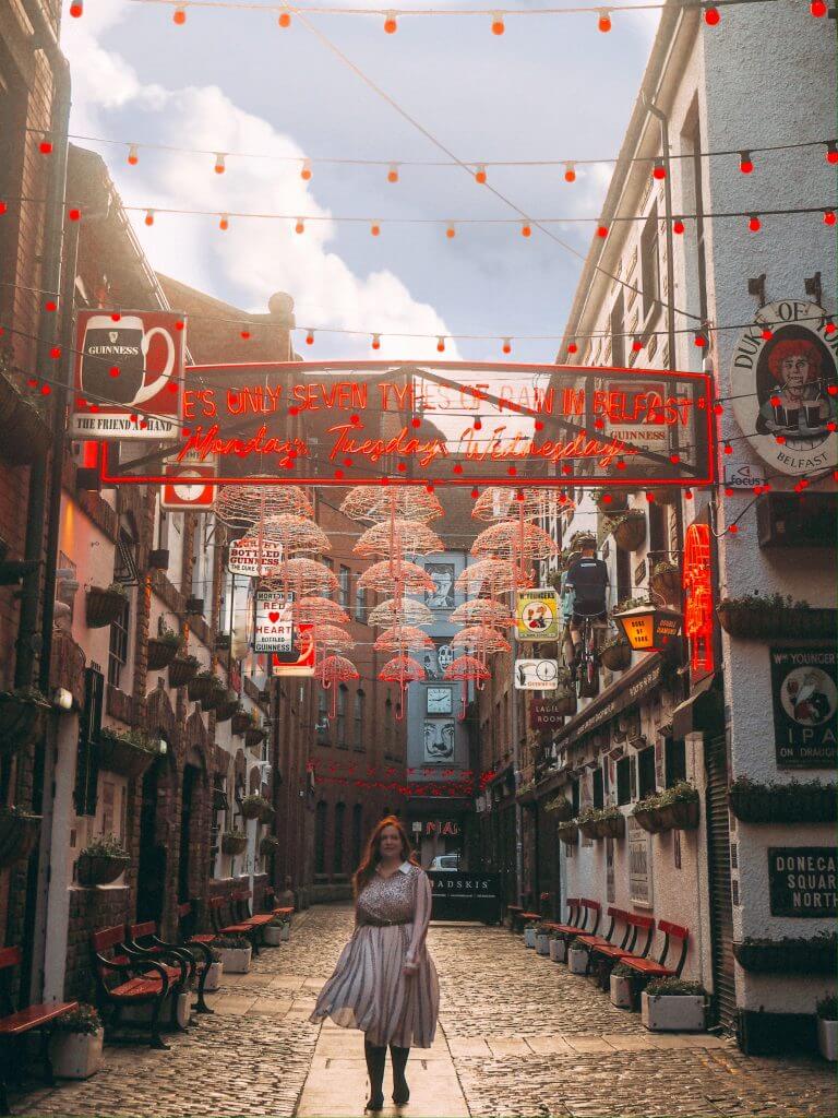 Woman in a white dress walking along the cobbled streets of the cathedral quarter in Belfast, beside the Duke of York pub an Instagram spot in Belfast