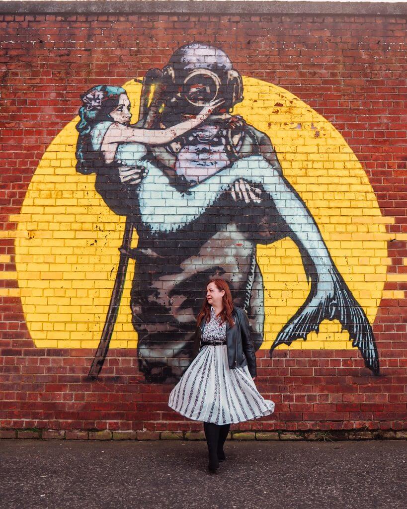 Woman wearing a leather jacket and white dress standing infront of a mural of a deep sea diver carrying a mermaid.