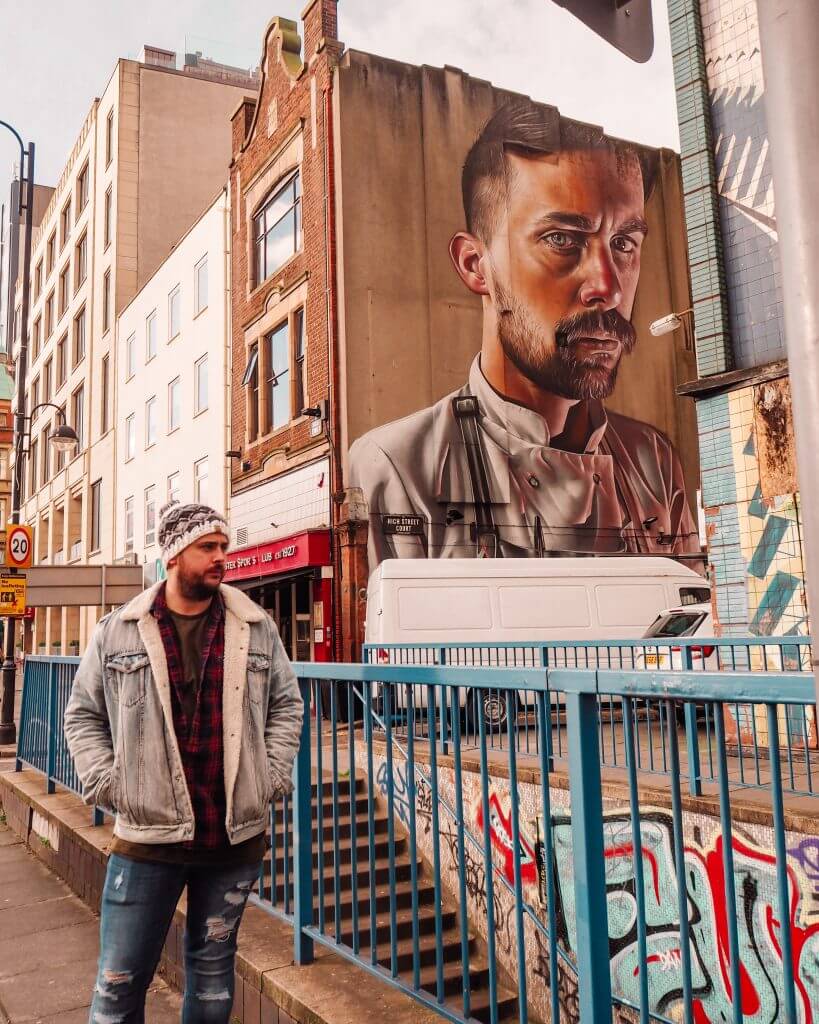 Man in a fleece lined denim jacket standing in front of The Chef by Smug One street art in Belfast city