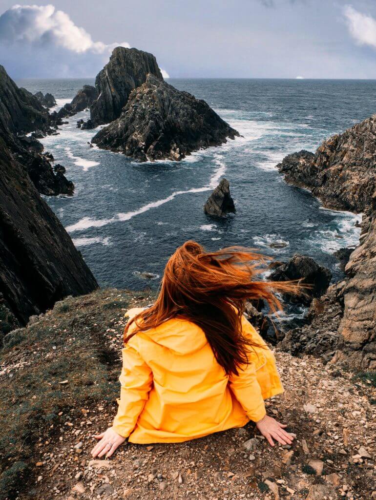 Woman wearing a yellow raincoat sitting at the cliff edge at Malin Head in County Donegal Ireland