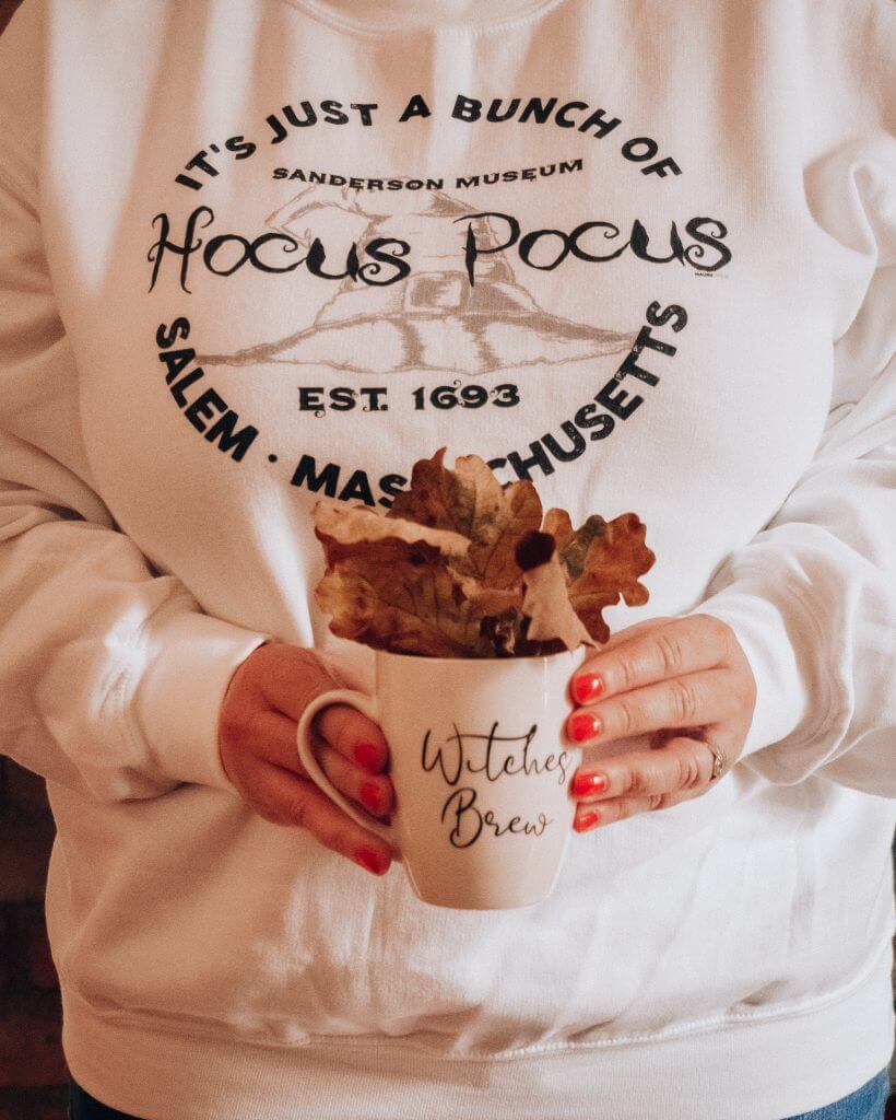 Woman holding a cup that say witches brew with fall leaves and wearing a hocus pocus sweater for an Instagram photo