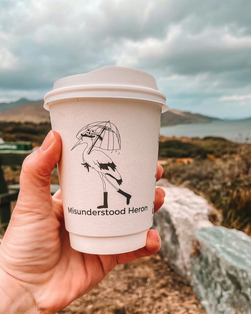 Hand holding a coffee cup from Misunderstood Heron over views of Connemara Ireland