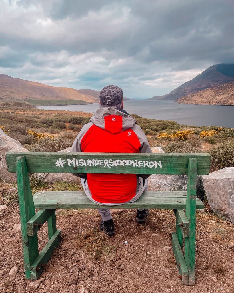Man in a red rain jacket and baseball cap sitting on a green bench at the Misunderstood Heron food truck with views over Killary harbour.