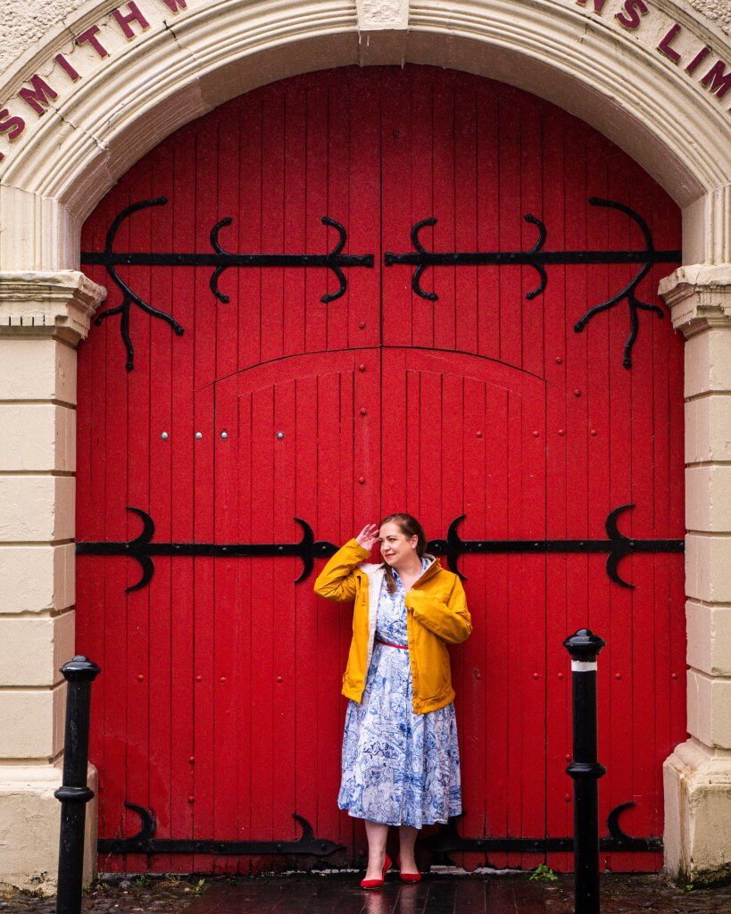 Woman in a yellow raincoat standing in front of the red gates of the Smithwick's factory in Kilkenny Ireland one of the many things to do on a weekend in Kilkenny