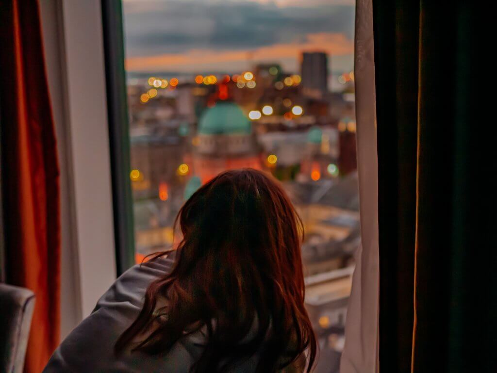 Woman looking from her hotel window at Grand Central Belfast at Belfast city at sunset