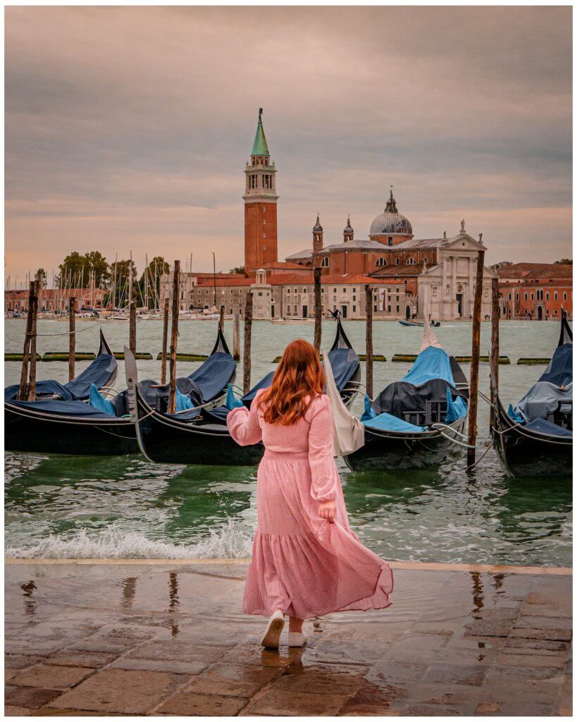 Woman in a pink dress splashing in the waves of the Grand Canal in Venice with a row of gondolas in the background.