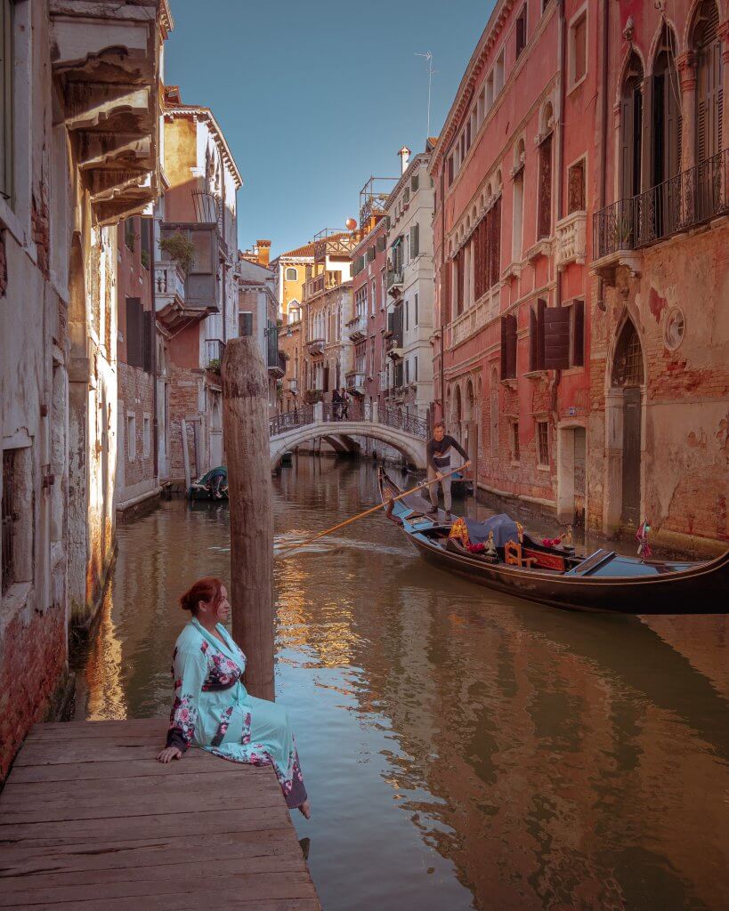 Woman sitting at a hidden pier in San Moise Venice as a gondola goes past. A hidden Instagram Spot in Venice.