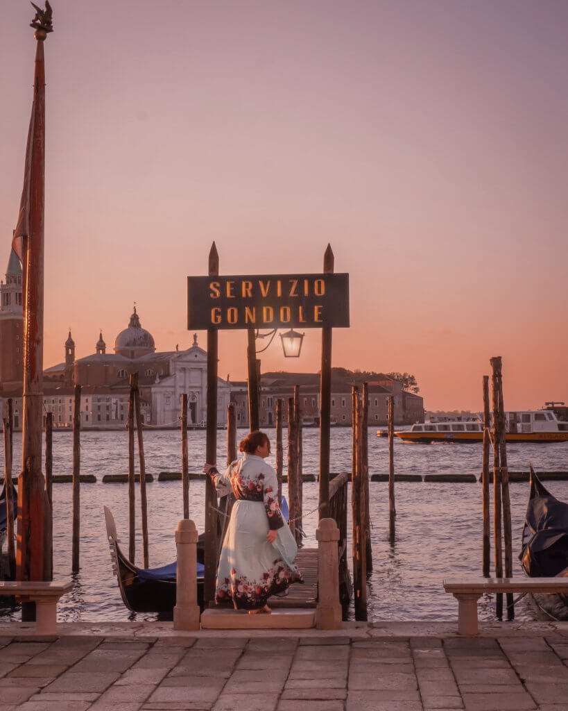 Woman in a long blue floral dress standing at a sign for gondolas on the Grand Canal in Venice Italy at sunset. Best Instagram spots in Venice.