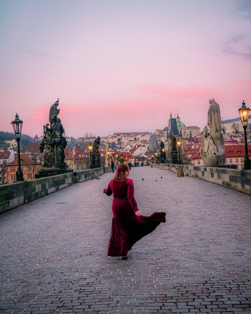 Woman in a long red velvet dress walking along Charles Bridge at sunrise
