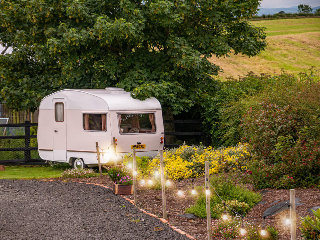 Caravan in a garden of flowers with string lights hanging along the path.