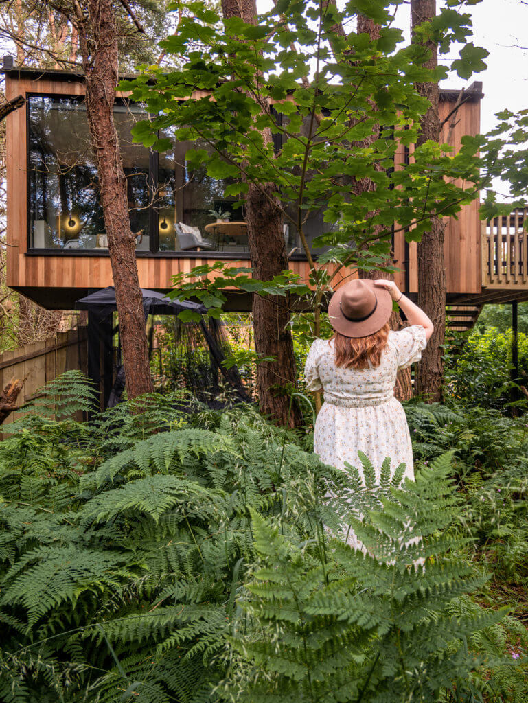Woman in a floral dress and a brown hat looking up at her luxury treehouse in Ireland