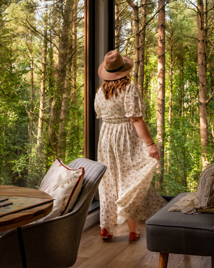 Woman wearing a floral summer dress and hat looking out at the trees from her romantic treehouse Ireland