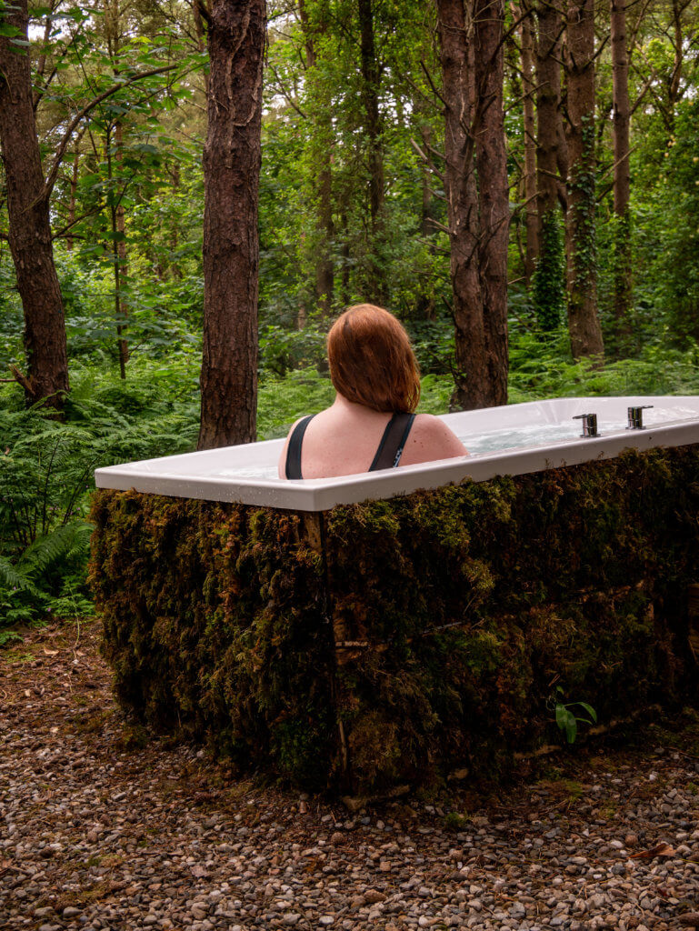 Woman sitting in a moss covered outdoor whirlpool bath in the forest at Burrenmore Nest Ireland