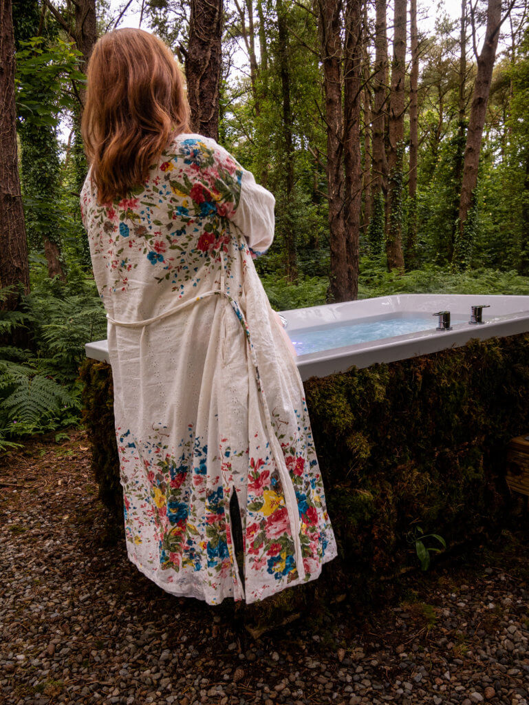 Woman wearing a floral robe sitting on a moss covered outdoor whirlpool bath at a luxury glamping accommodation in Ireland