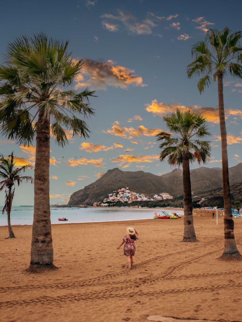 Woman wearing a hat and a floral dress walking in the sand on a beach with palm trees at Playa de Las Teresitas while exploring Tenerife by car.