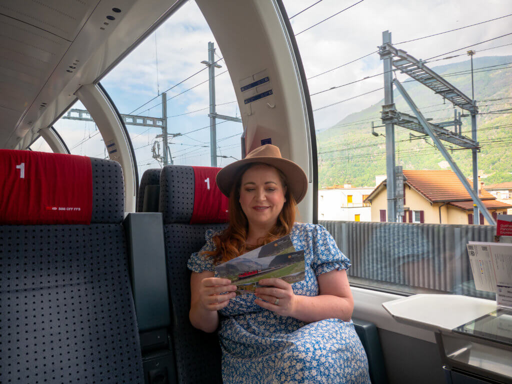 Nicola wearing a hat and a blue dress reading postcards on the Gotthard Panorama Express