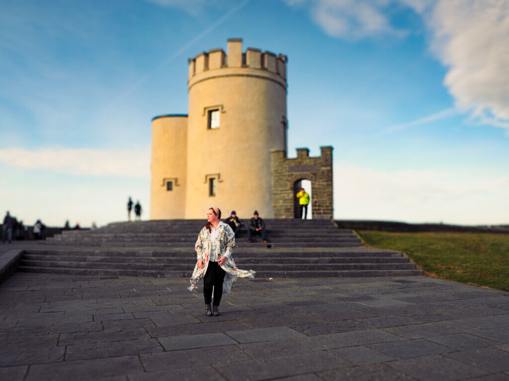 Nicola Lavin, top 10 travel influencer in Ireland is walking in front of the iconic O'Brien's Tower, standing tall at the highest point of the Cliffs of Moher. She is wearing a pink headband, black jeans and a long, floral, flowing kimono.