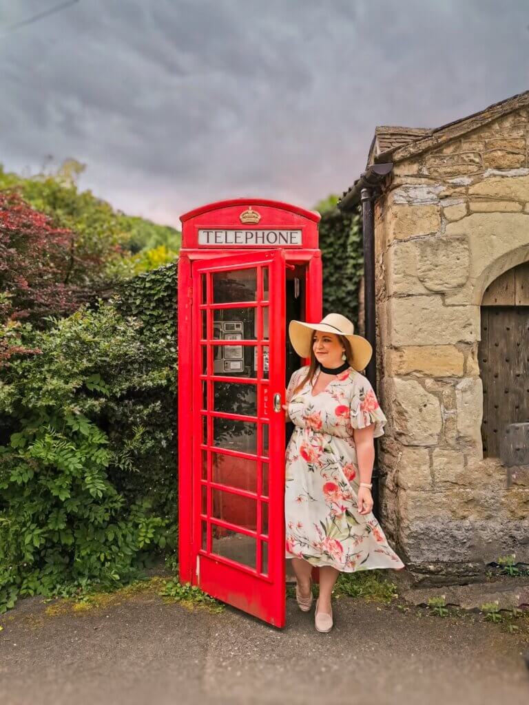 A woman in a floral dress wears a floppy sun hat and stands at a red telephone box in the idyllic village of Castle Combe in the Cotswolds.