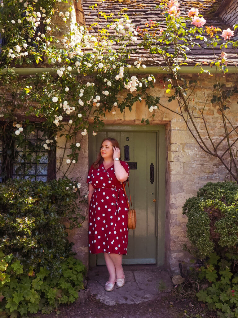 Nicola Lavin, an Irish travel blogger, stands against a quaint thatched-roof cottage framed by colourful flowers in the Cotswolds.