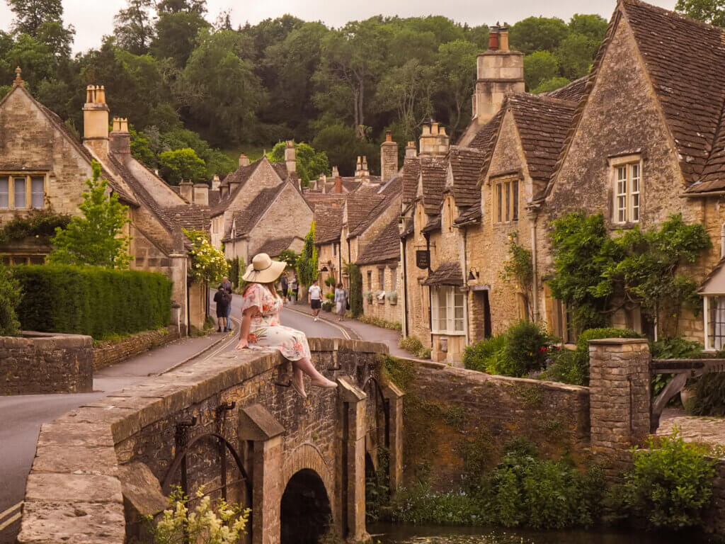 Nicola Lavin, an Irish travel blogger sits on the bridge of the fairytale village of Castle Combe in the Cotswolds. She is wearing a floral summer dress and a floppy hat.
